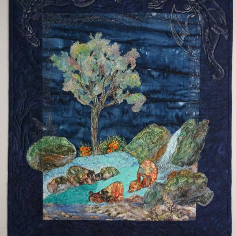 Karin Tauber - Dreaming of Dragons (a quilt landscape showing a tree, river and rocks. Stitched dragons fly around the dark indigo border.)